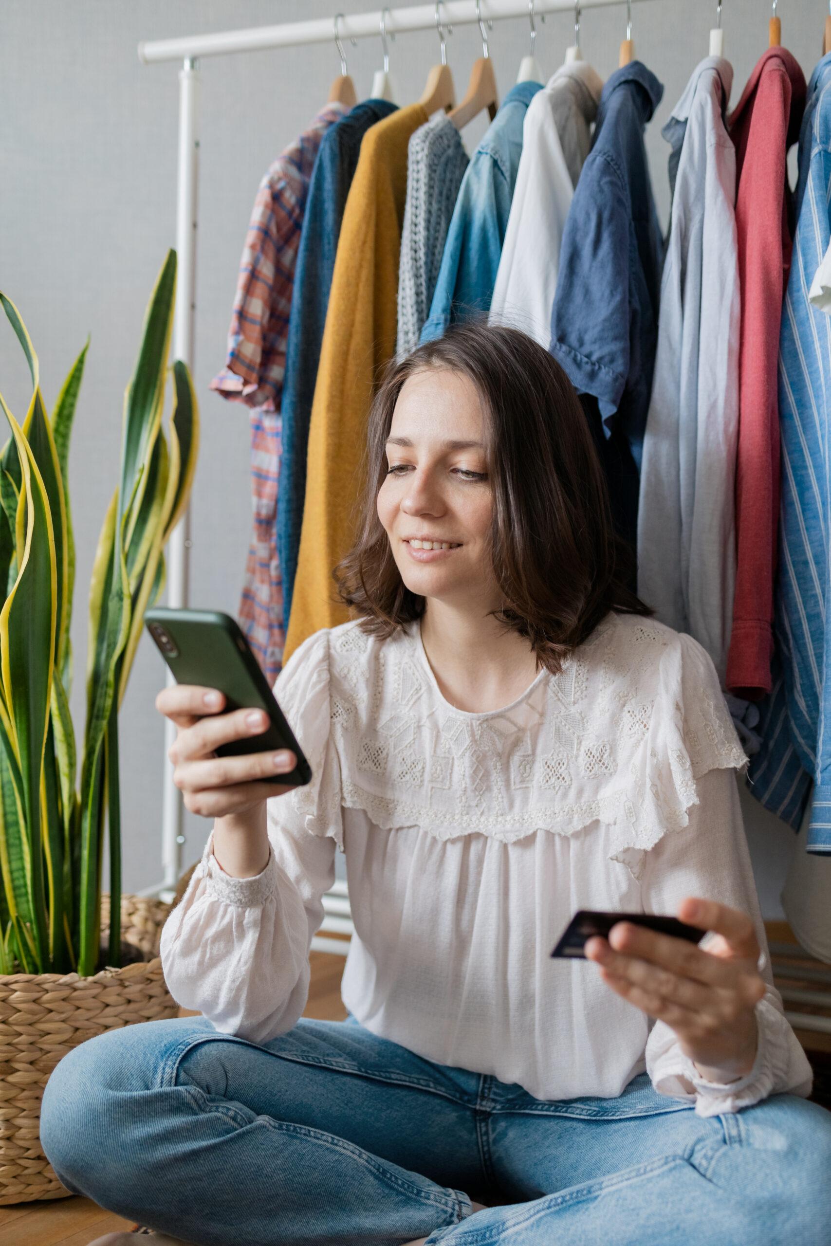 Beautiful European woman sitting in her Wardrobe, among the clothes and makes a purchase online, a woman with a phone and a bank card to order a new dress